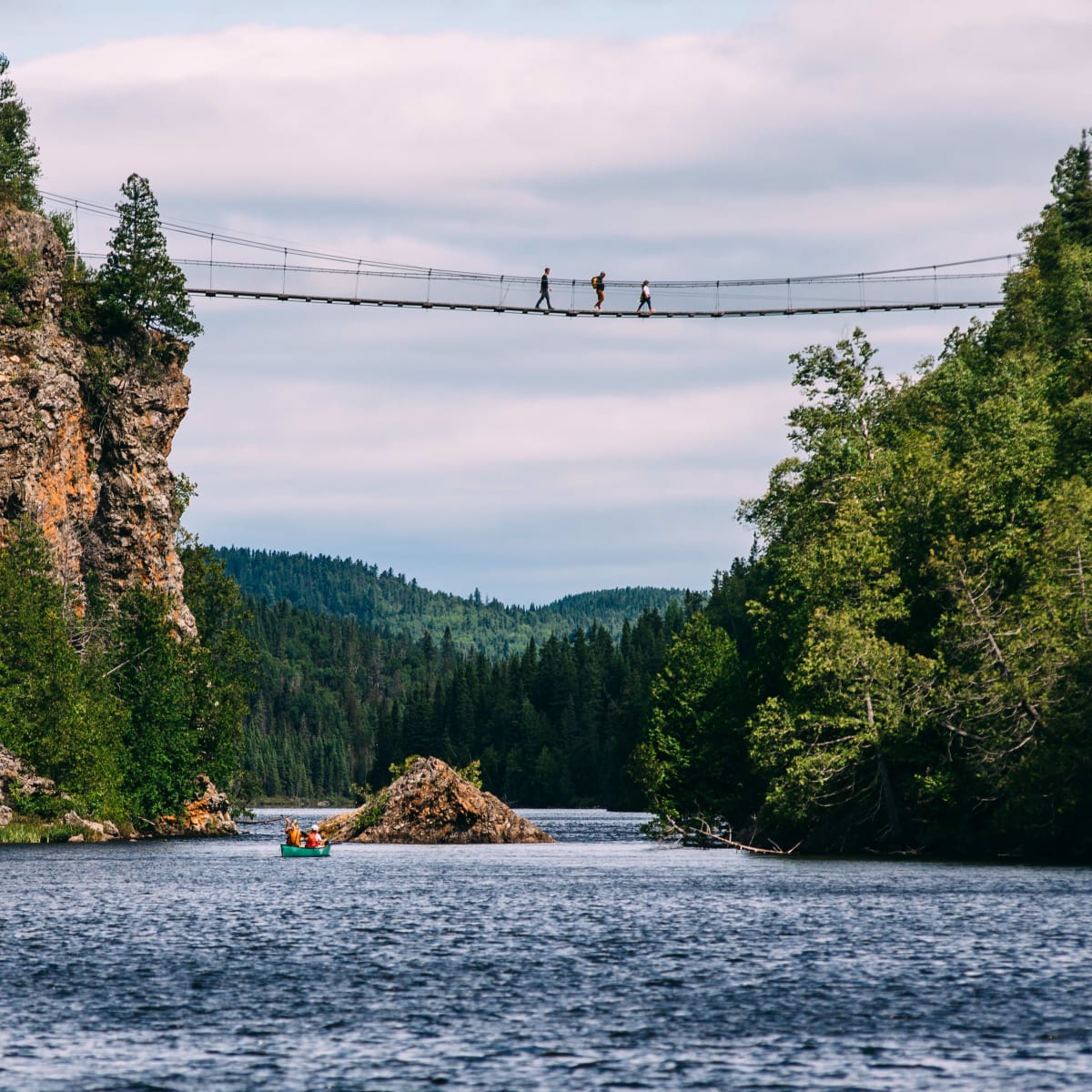 Pont suspendu au-dessus d'un plan d'eau; il y a des personnes sur le pont ainsi qu'une embarcation sur l'eau.