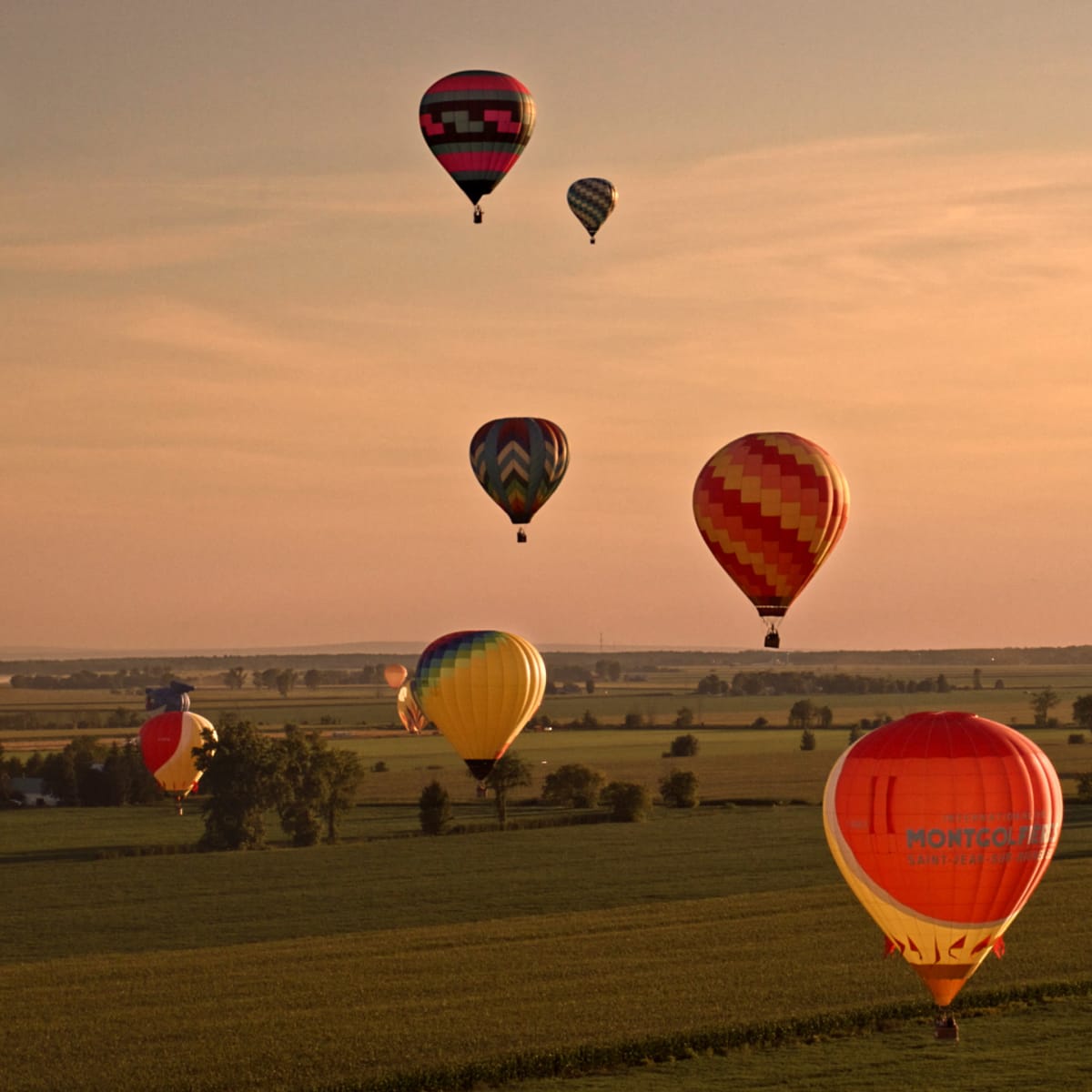 Une dizaine de montgolfière dans un ciel orangé au-dessus de champs s'étendant vers l'horizon.