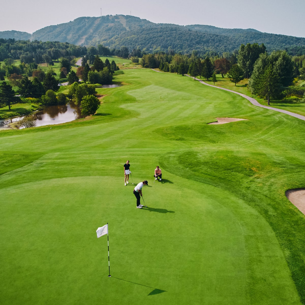 Trois personnes sur un vert au Golf Château-Bromont, avec une montagne en arrière-plan.