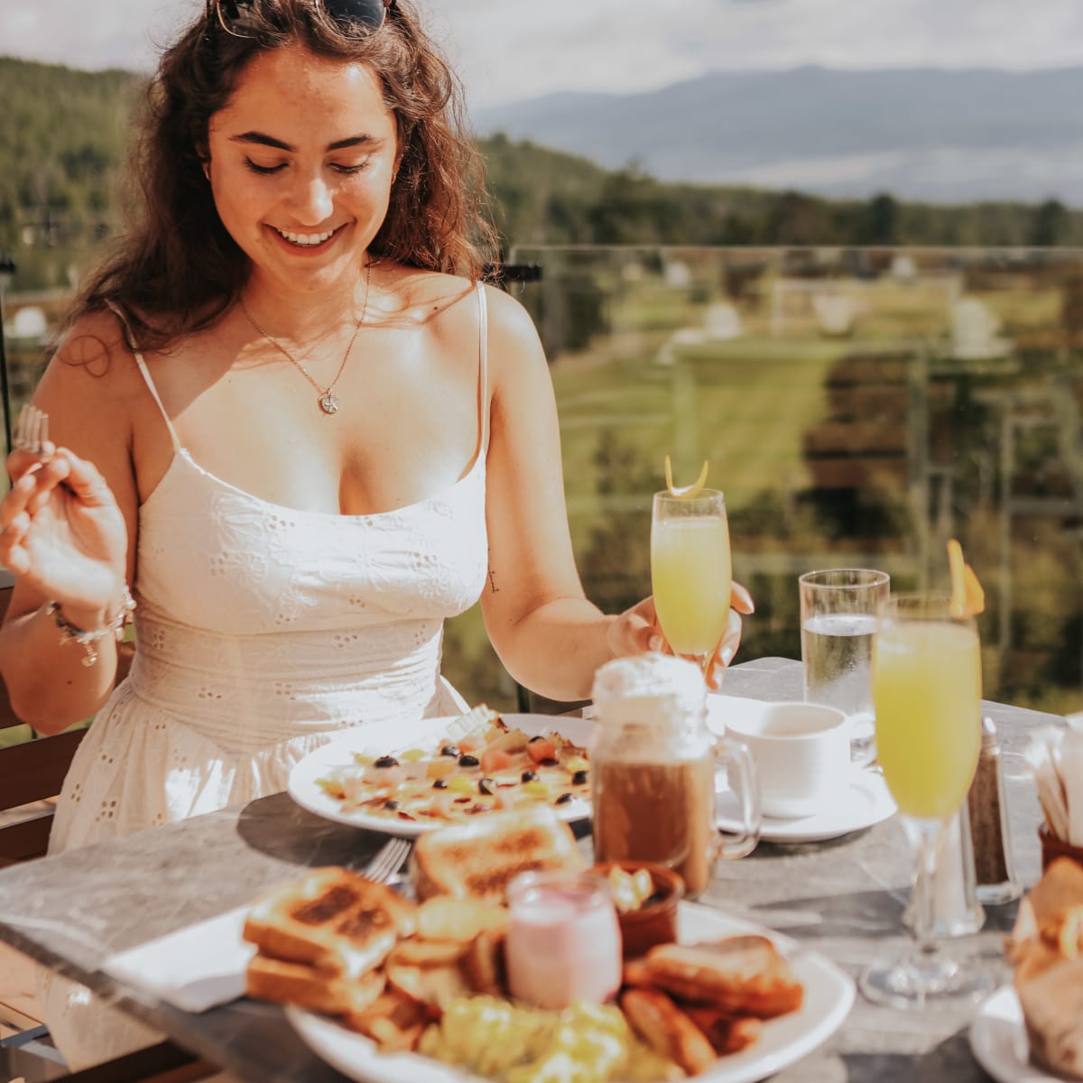 Femme souriante sur une terrasse avec des assiettes bien remplies sur la table devant elle.