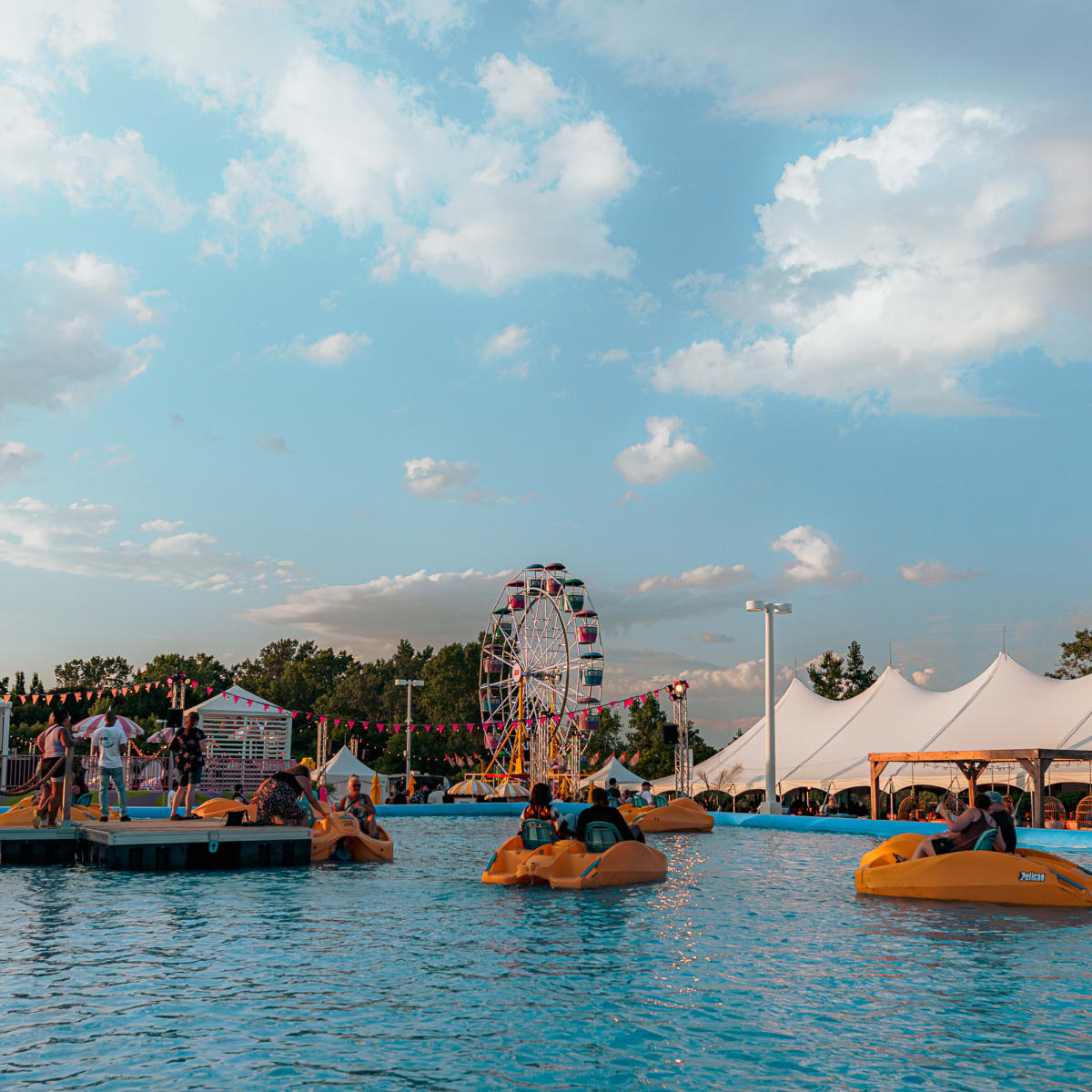 Festival Parasol - Des personnes font du pédalo dans une grande piscine extérieure, avec une grande roue et des tentes blanches en arrière plan