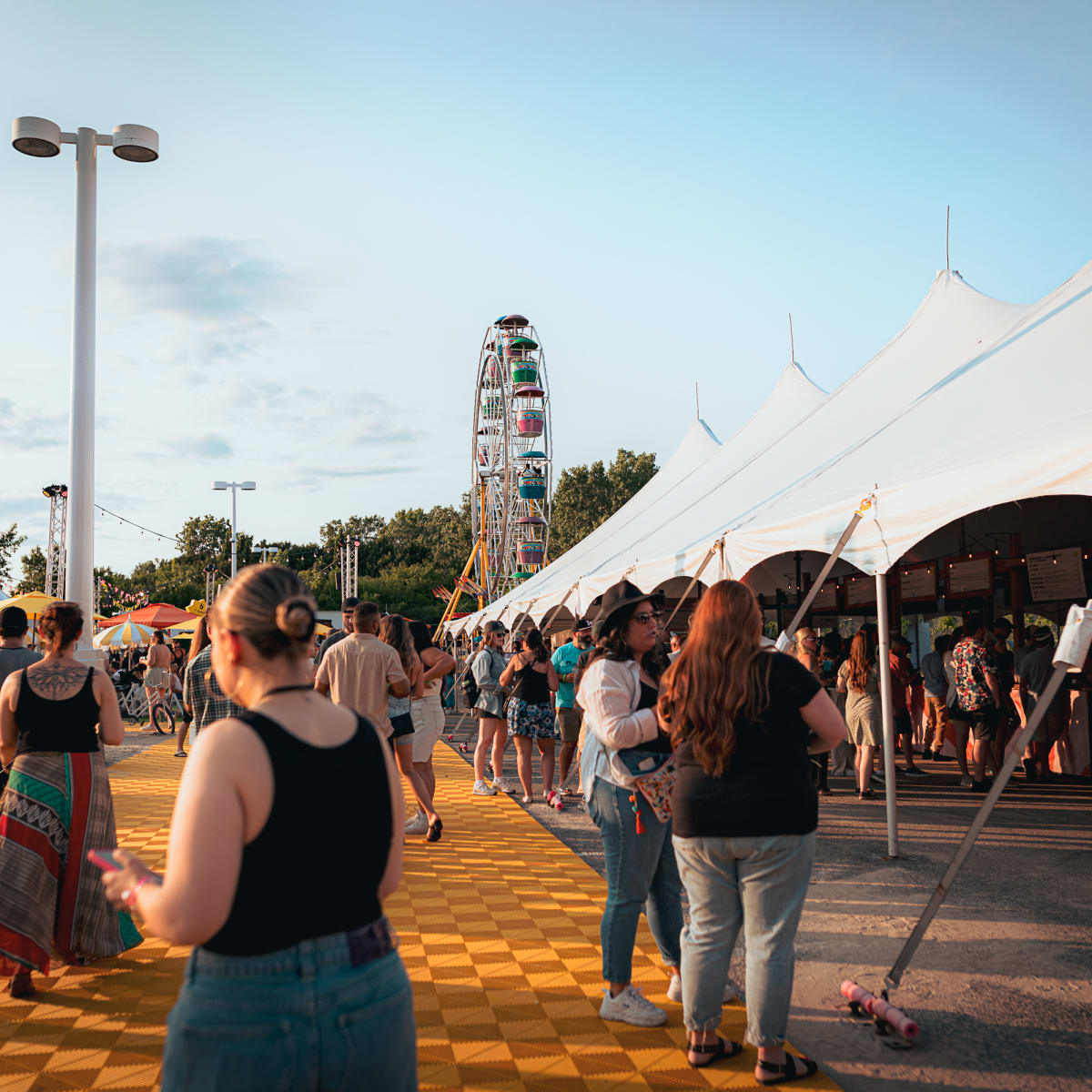 Festival Parasol : Des personnes sont éparpillés sur le site. Un grand chapiteau blanc se trouve à droite, avec une grande roue en arrière-plan.