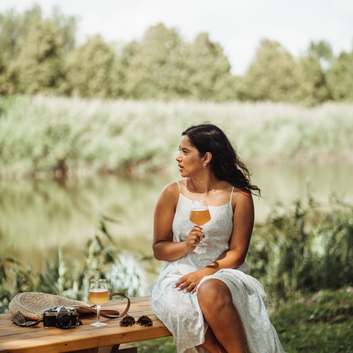 Femme assise sur une table à pique-nique qui déguste un verre de bière dans un paysage naturel.