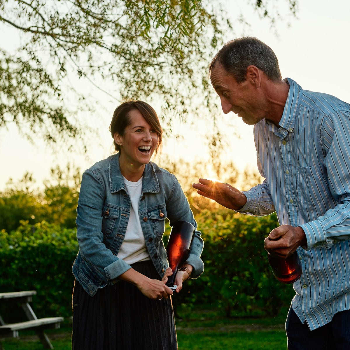 Au coucher de soleil, une femme et un homme souriants tiennent chacun une bouteille de vin à la main.