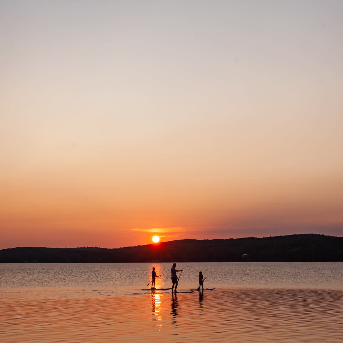 Trois personnes en planche à pagaie sur un grand plan d'eau, devant un soleil couchant orangé.
