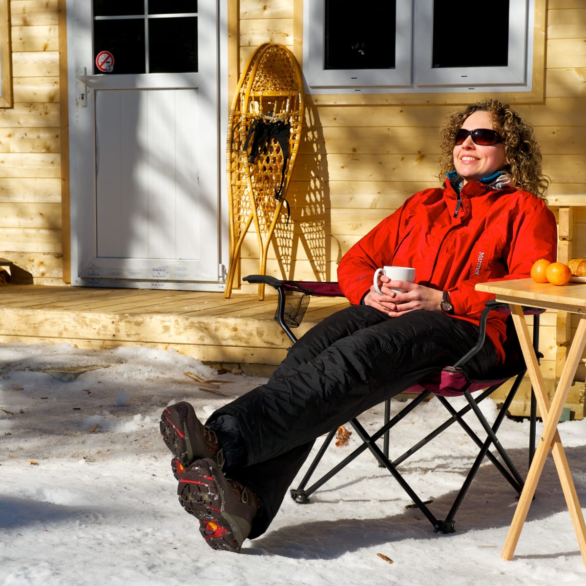 Madame assise dehors l'hiver au soleil avec un déjeuner et des raquettes.