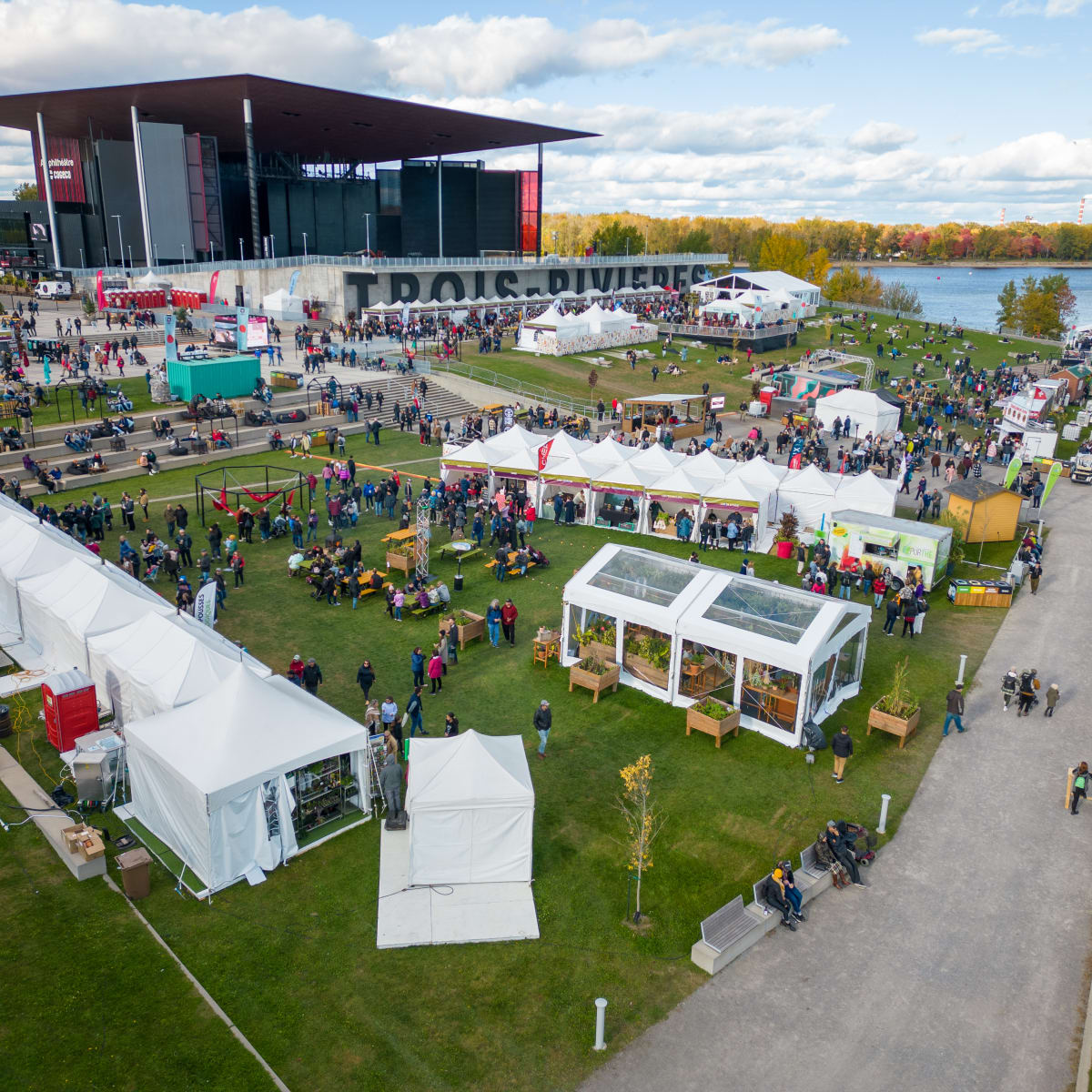 L'Amphithéatre Cogeco à Trois-Rivières au bord du fleuve avec des kiosques et spectateurs sur l'esplanade en gazon.