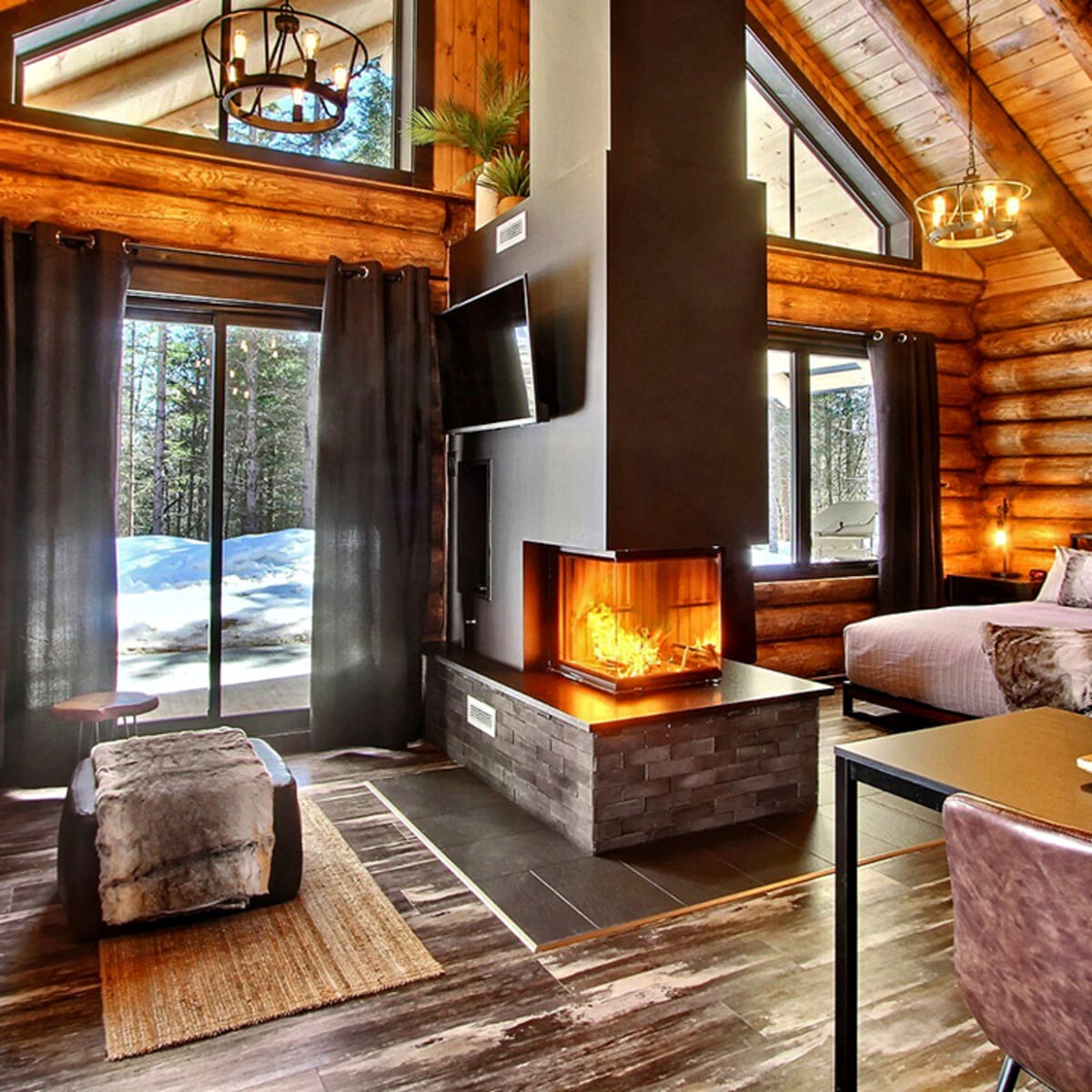 Interior of a log cabin with a central fireplace and large windows.