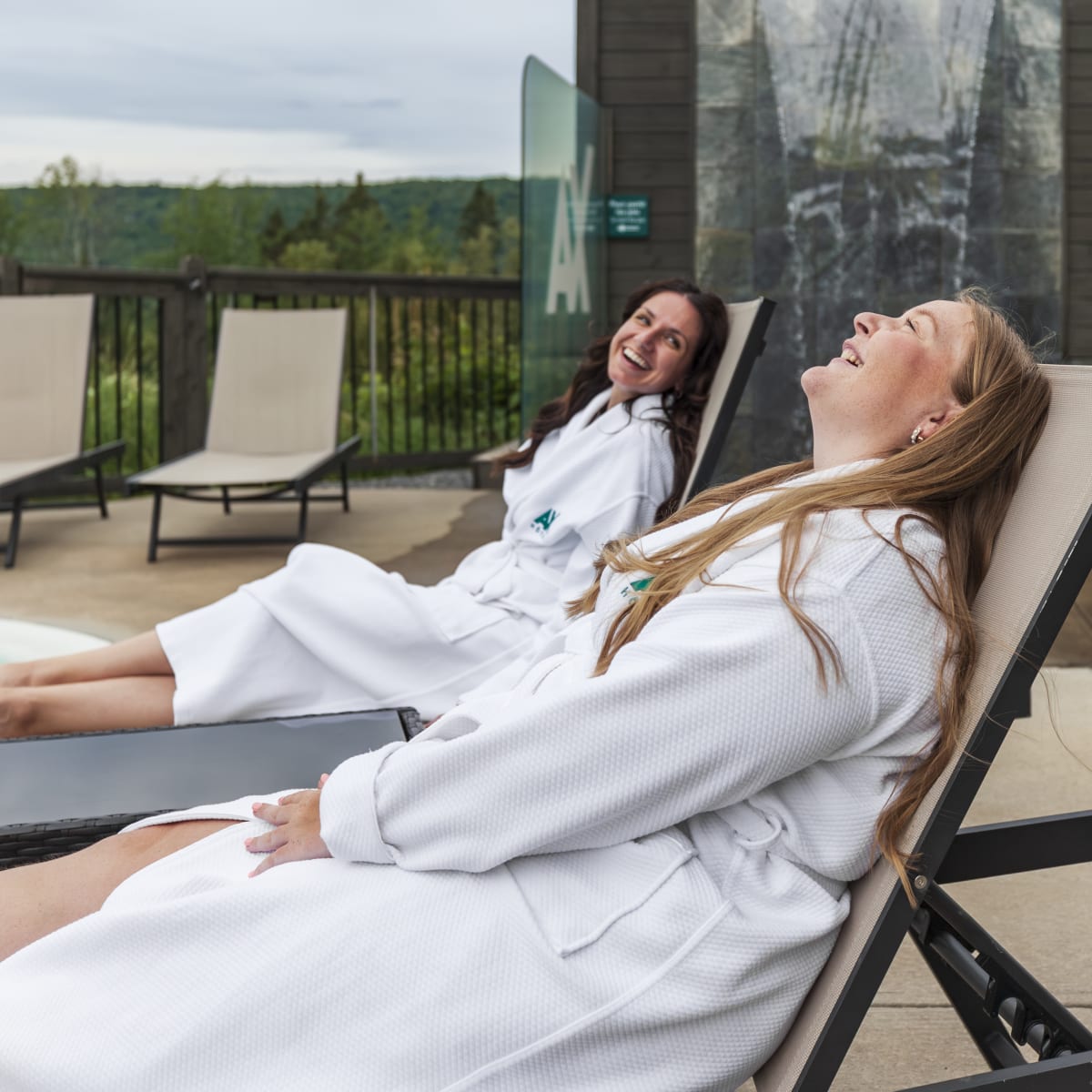 Two women in bathrobes laughing while lying on lounge chairs.