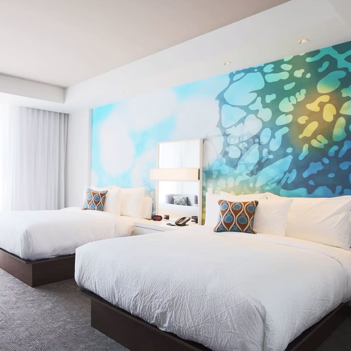Chambre d’hôtel illuminée de lumière naturelle, avec deux grands lits, literie blanche et mur d’accent bleu.