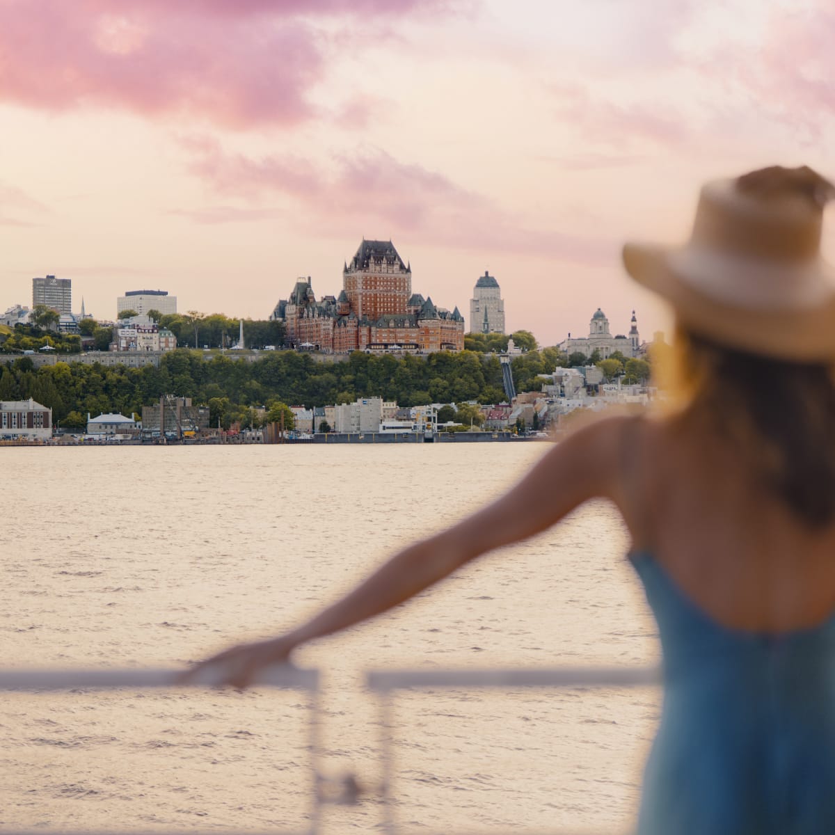 Femme en robe bleue avec un grand chapeau observant le Château Frontenac depuis le fleuve.