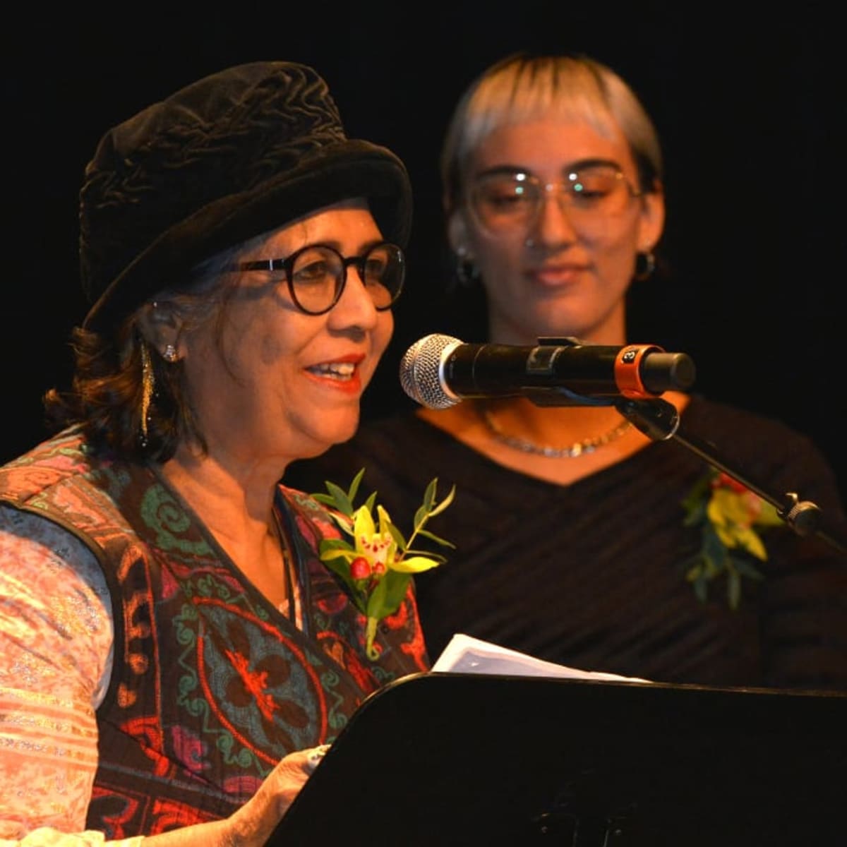 A woman wearing a hat recites a poem in front of a microphone and podium and a young woman stands beside her.