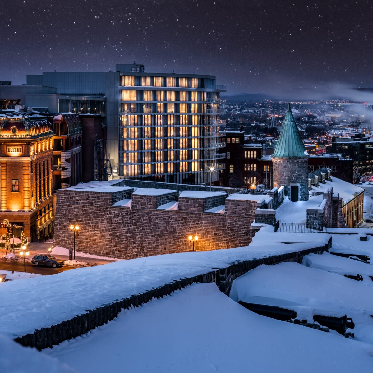The hotel’s modern building amidst historic buildings under snow; the sky is starry and the city is illuminated in the distance.