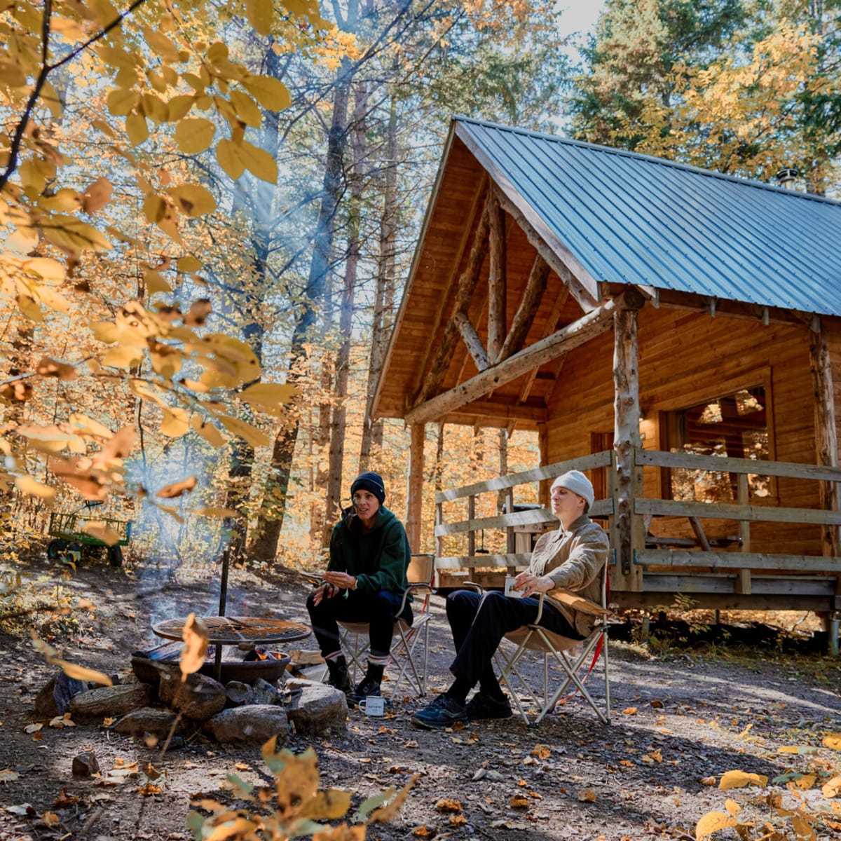 Deux personnes assises autour d’un feu de camp devant un chalet en bois dans une forêt à l’automne.