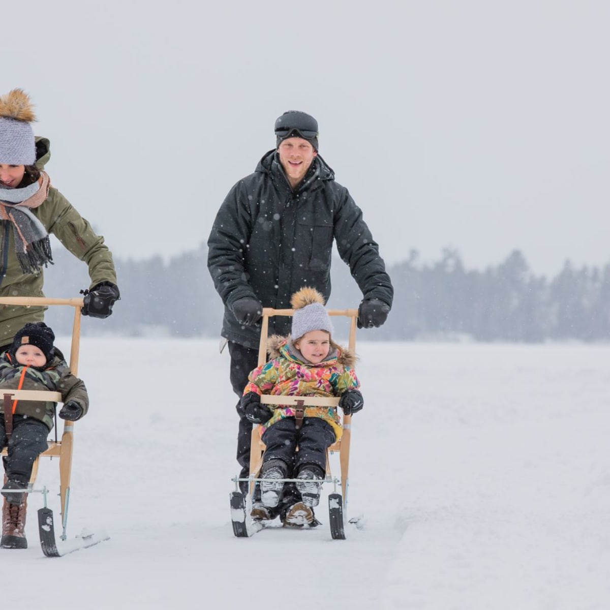 Two parents pushing two kids in snow scooters.