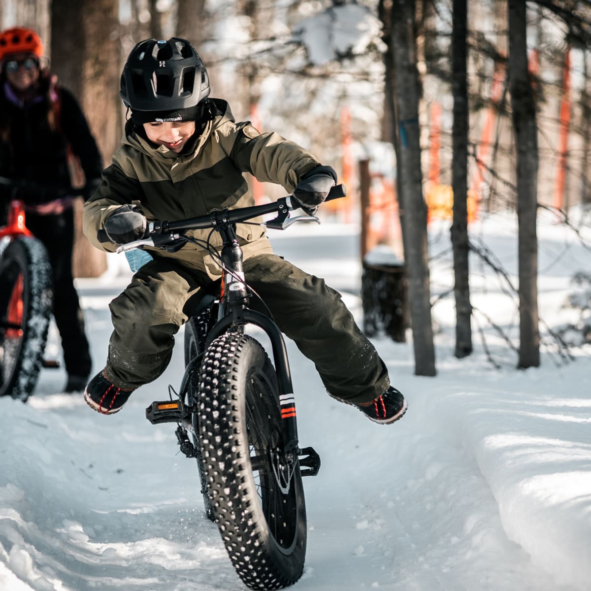 Jeune garçon en fatbike dans une forêt enneigée en avant-plan, avec une autre personne plus loin derrière.
