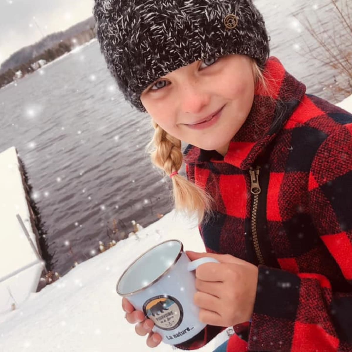 Young girl with a hat and plaid shirt by the water in the winter holding a mug