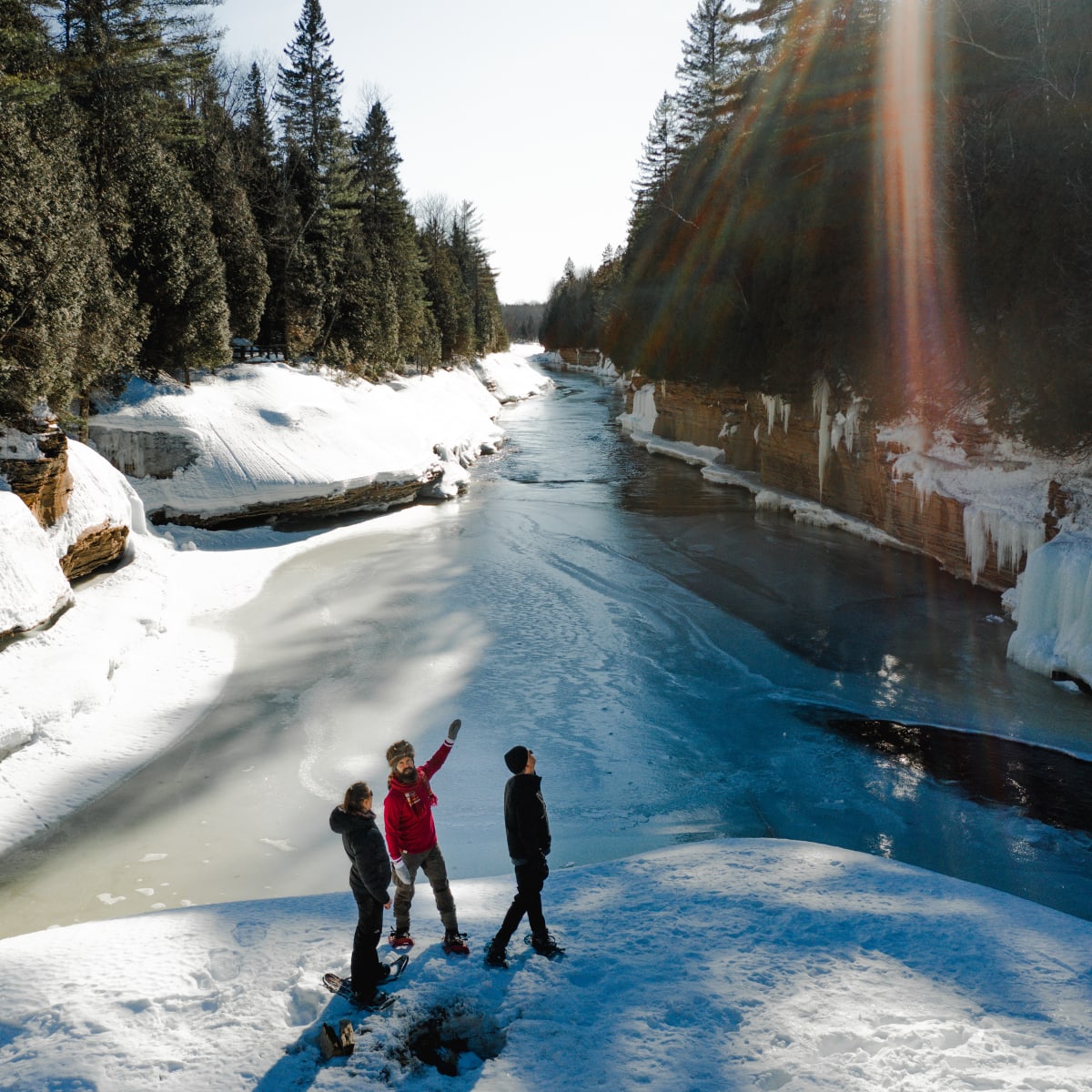 3 people wearing snowshoes crossing a river in the forest in winter.
