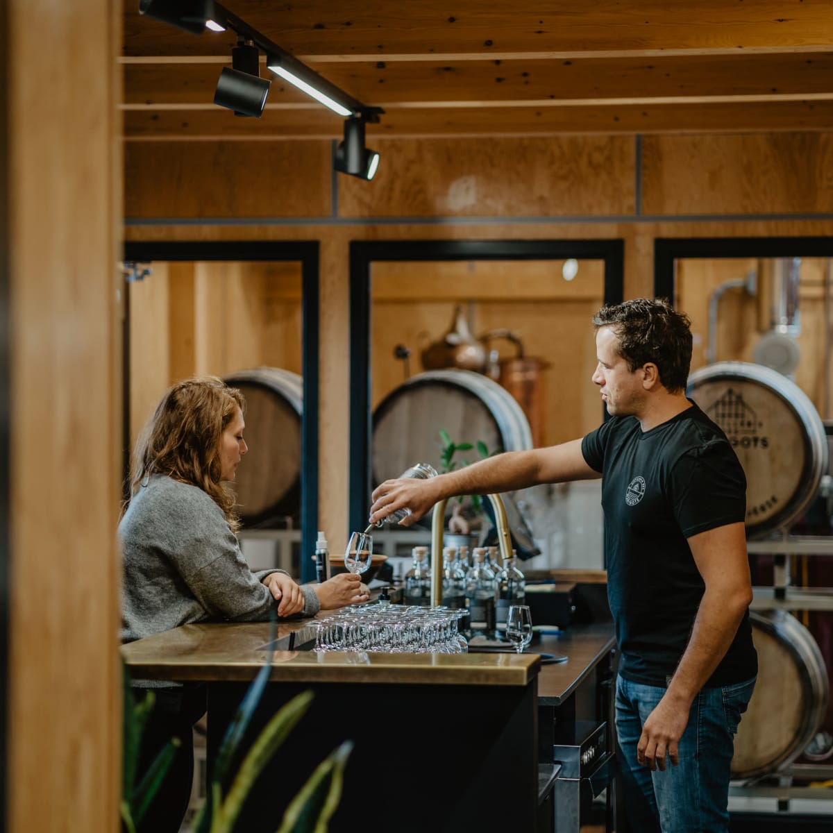A man serving a drink to a woman in a wood-paneled tasting room, with stacked barrels in the background.