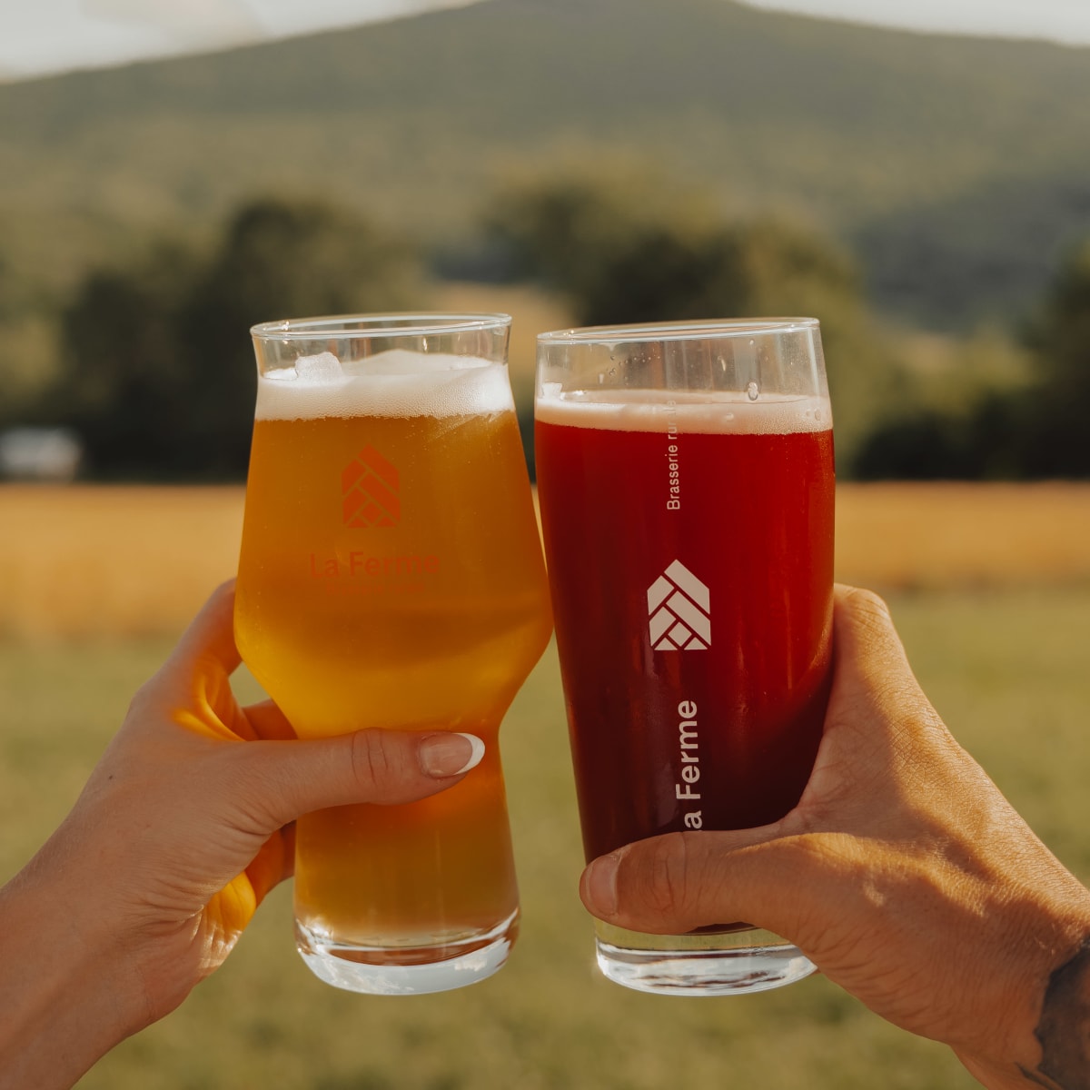 Two glasses of beer held by people outside the frame in front of a mountainous landscape.