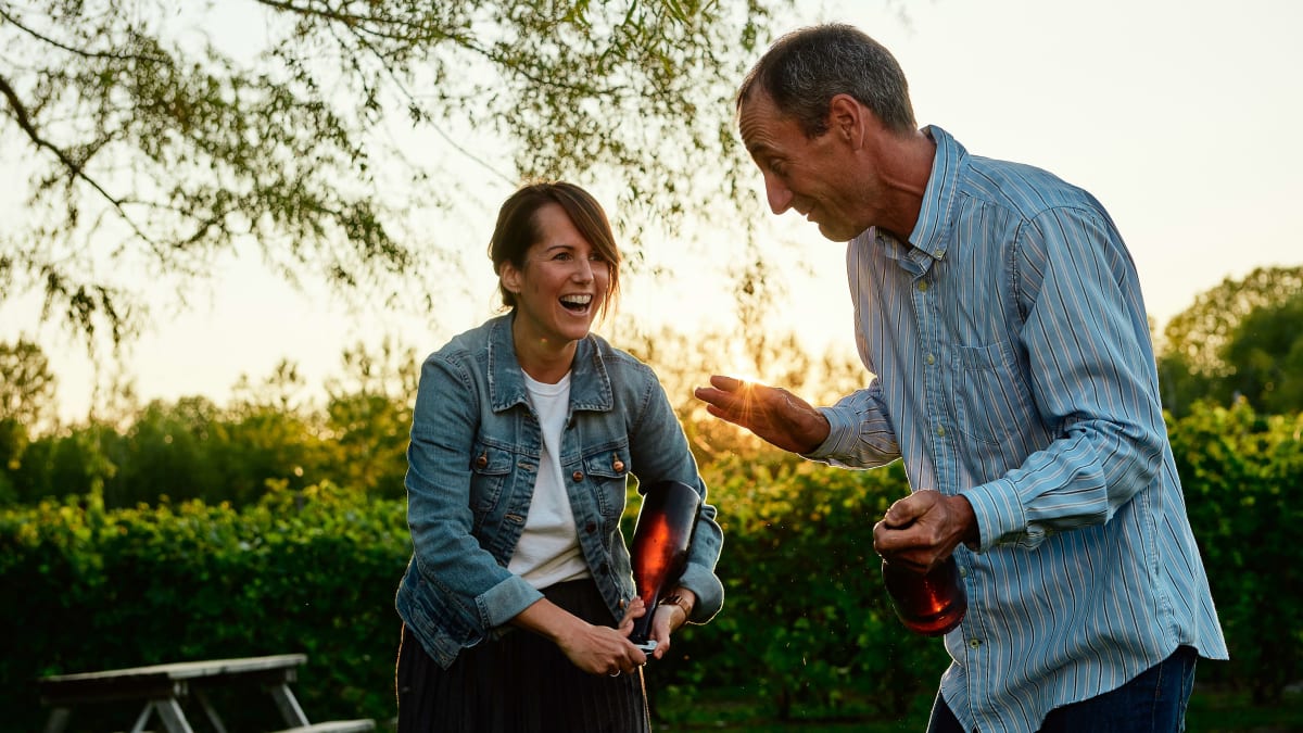 Au coucher de soleil, une femme et un homme souriants tiennent chacun une bouteille de vin à la main.