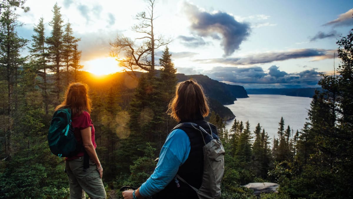 Parc national du Fjord-du-Saguenay - Deux femmes en haut d'une montagne, la forêt et le couché de soleil en arrière-plan.