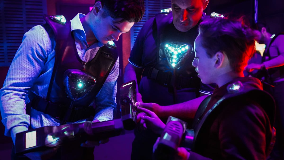 Three people of varying ages grouped together in a laser tag arena, equipped with glowing vests and laser guns.