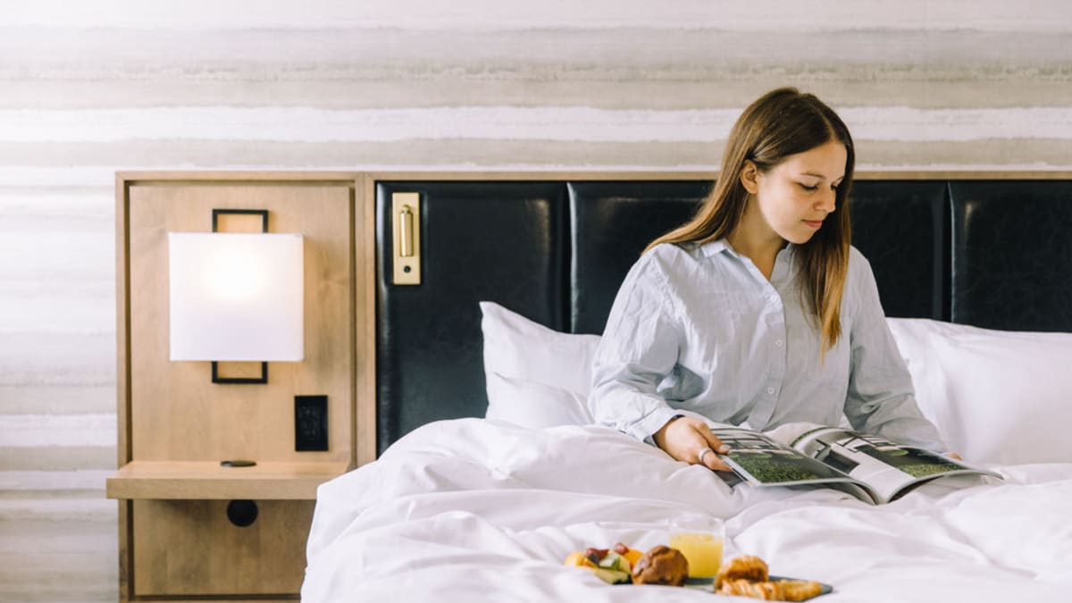 Une femme en pyjama est assise sur un lit. Elle lit un magazine et il y a un plateau déjeuner sur la douillette.