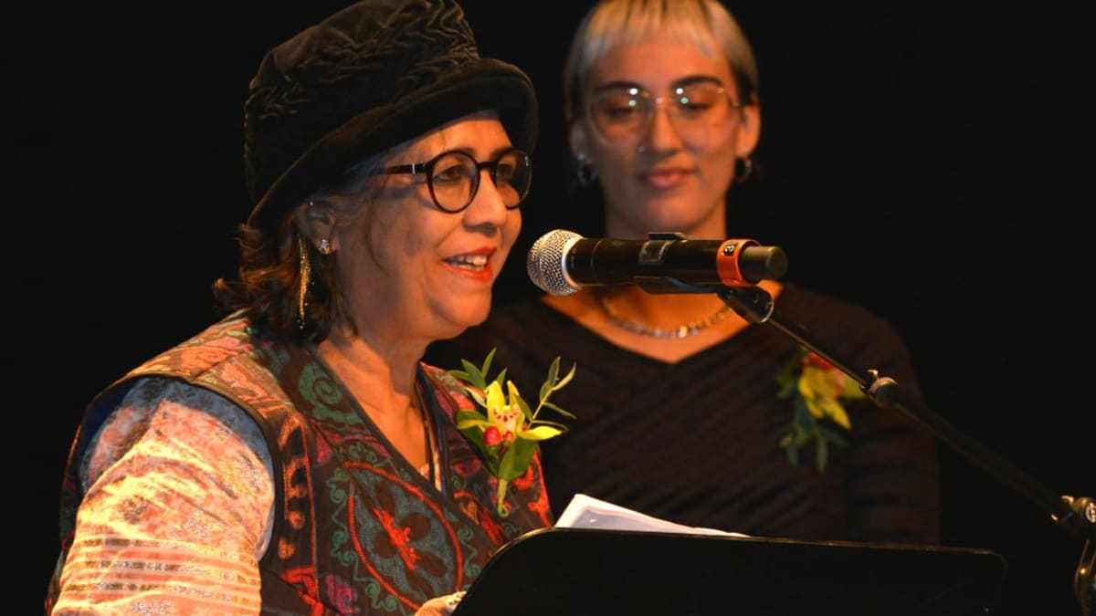 A woman wearing a hat recites a poem in front of a microphone and podium and a young woman stands beside her.