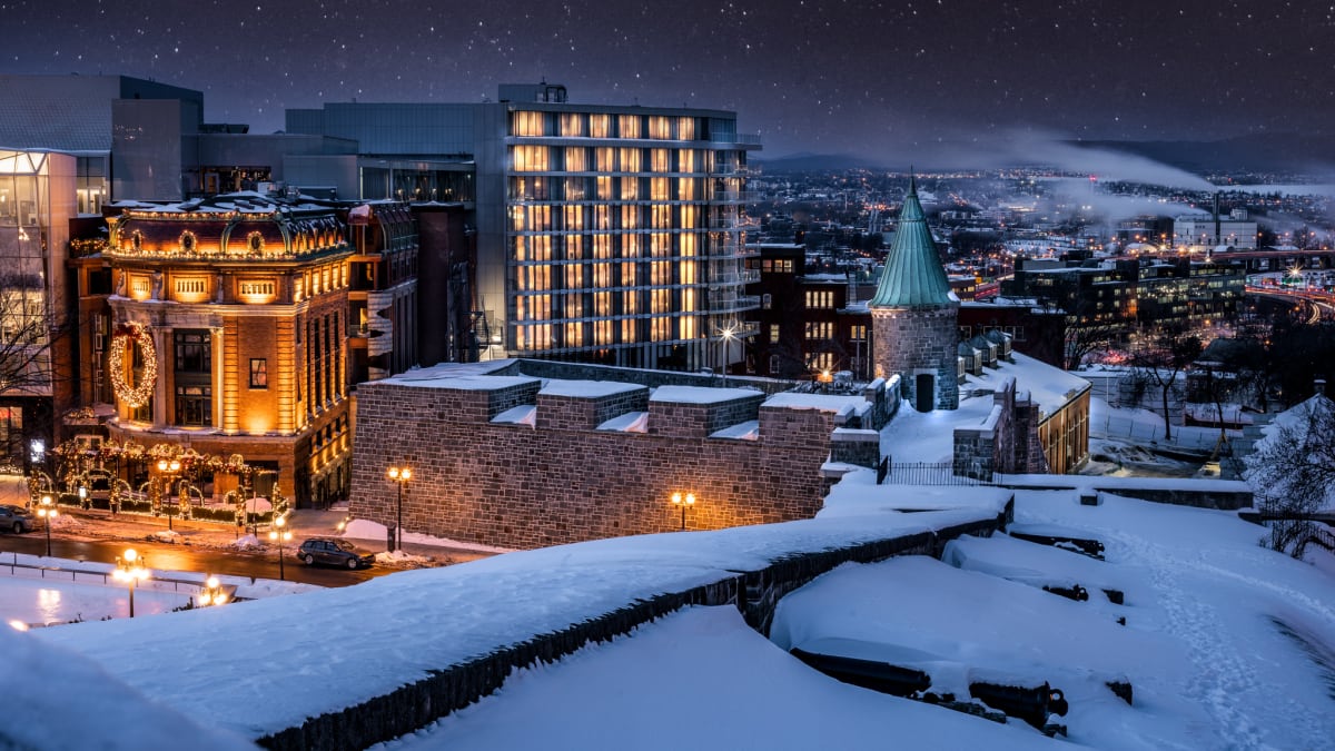 L’édifice moderne de l’hôtel parmi des bâtiments historiques sous la neige; le ciel est étoilé et la ville est illuminée au loin.