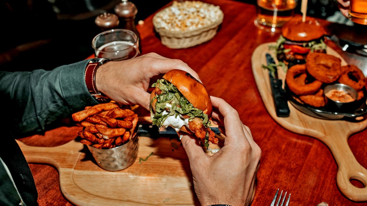Deux mains tenant un hamburger au-dessus d'une planche avec des frites sur une table de restaurant.