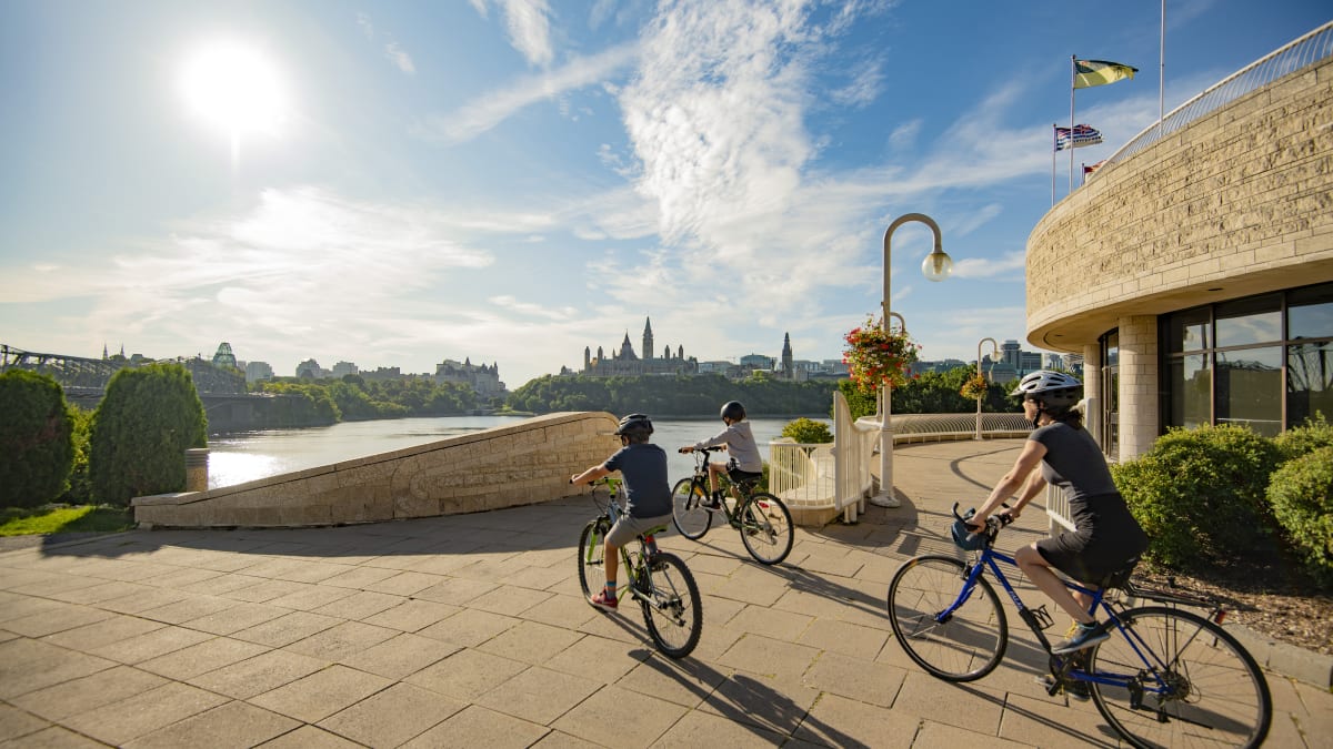 Mother and two sons on bikes next to the Canadian Museum of History in front of the Ottawa River.