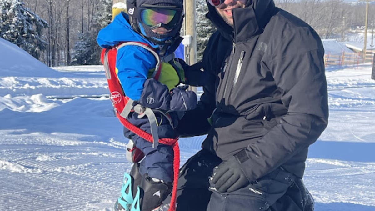 Un père avec son garçon, le bambin a une planche à neige aux pieds et le père est agenouillé devant lui.