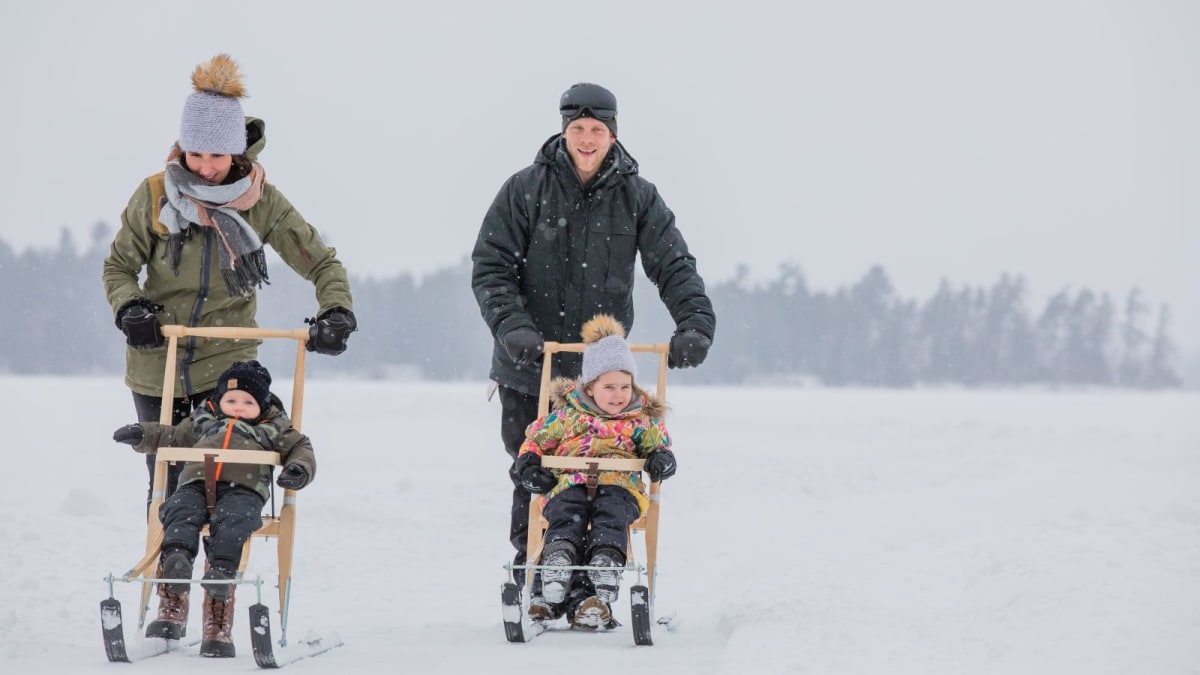 Two parents pushing two kids in snow scooters.