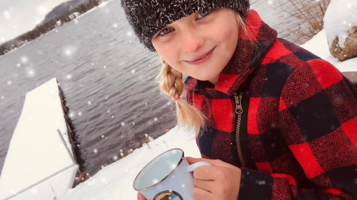 Young girl with a hat and plaid shirt by the water in the winter holding a mug