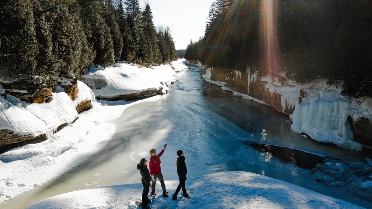 3 people wearing snowshoes crossing a river in the forest in winter.