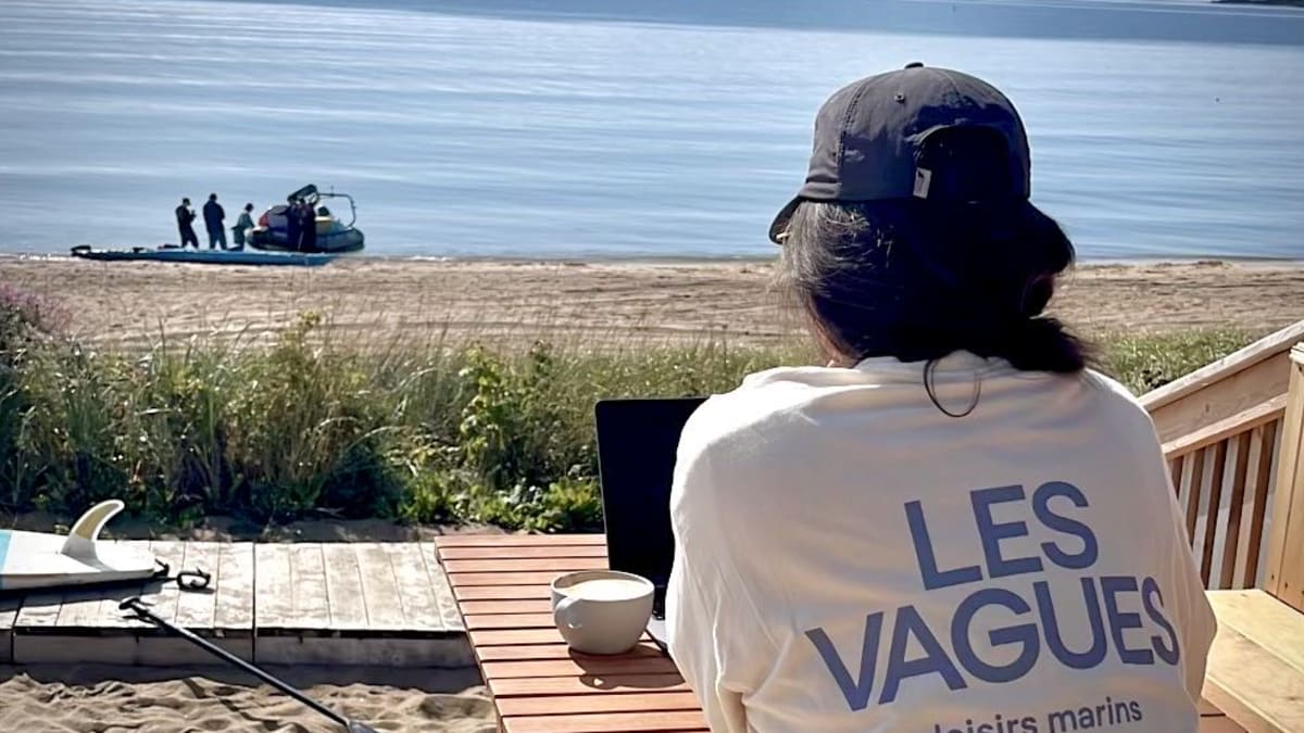 Woman wearing a Les Vagues T-shirt sitting at a café table by the water.