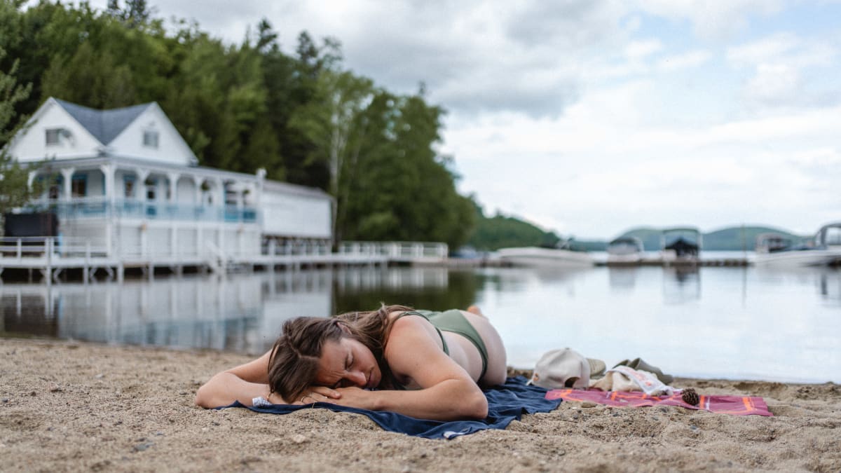 A woman lying on a beach; in the background, a body of water and a waterfront inn.