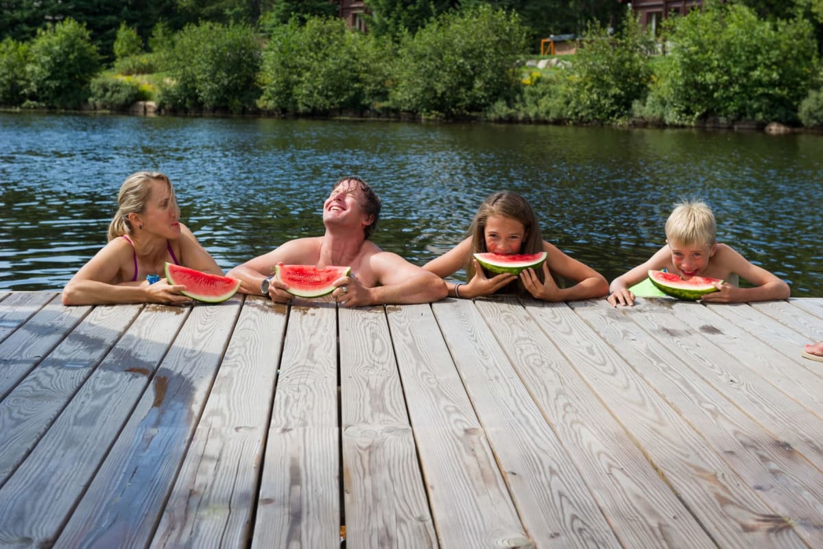 Quatre baigneurs dans l'eau, leurs bras posés sur un quai, en train de manger du melon d'eau.