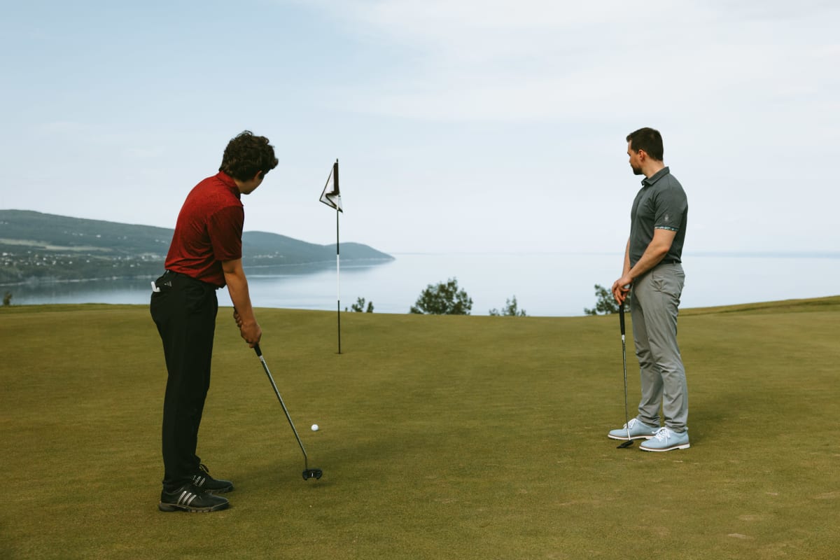 Jeune homme qui s'apprête à jouer un coup au Golf Manoir Richelieu, son père le regarde et le fleuve s'étend derrière eux.