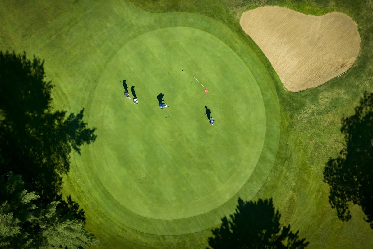 Vue aérienne d'un vert de golf bordé d'une fosse de sable; on y voit 4 golfeurs et leurs ombres.