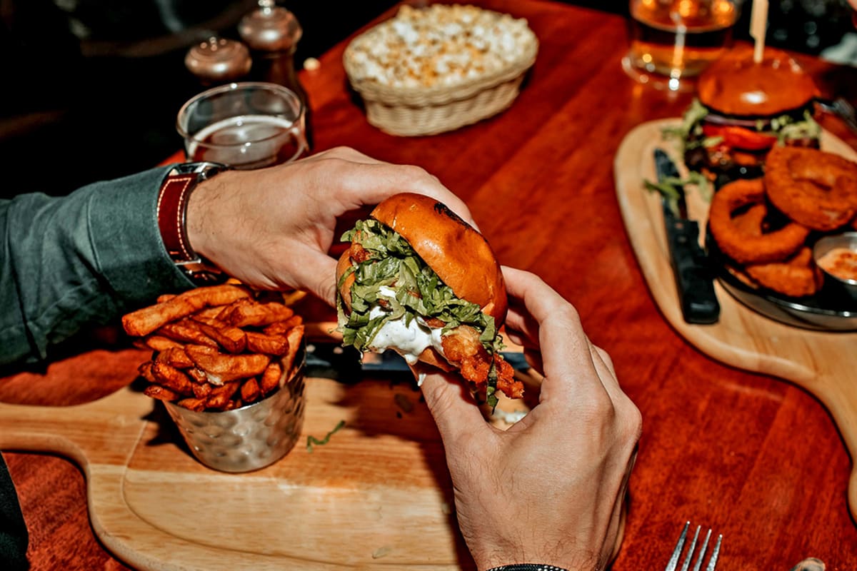 Deux mains tenant un hamburger au-dessus d'une planche avec des frites sur une table de restaurant.