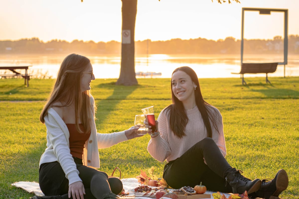 Two young women clink their glasses and share a picnic by the river,
