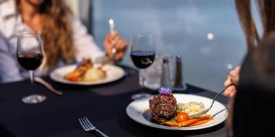 Deux personnes dégustent un repas gastronomique sur une table élégante avec une nappe noire, comprenant du vin rouge et des plats soigneusement présentés, le tout avec une vue sur l'eau en arrière-plan.