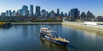 Un bateau de croisière naviguant sur une rivière, avec une vue panoramique sur une ville avec de hauts gratte-ciels en arrière-plan sous un ciel dégagé.