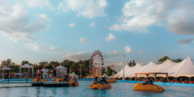 Festival Parasol - Des personnes font du pédalo dans une grande piscine extérieure, avec une grande roue et des tentes blanches en arrière plan
