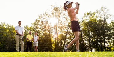 A woman plays golf, with three other individuals observing her in the background.