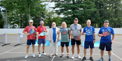 Jeux FADOQ provinciaux - On a pickleball court, there are six men wearing gold medals around their necks, and one man is dressed in a suit.