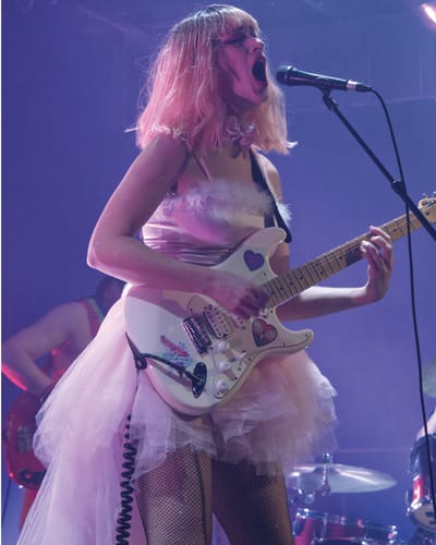 CEM - Une femme vêtue d'une robe rose chante et joue de la guitare électrique