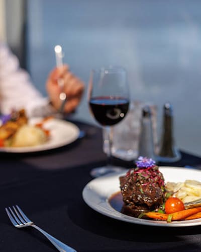 Deux personnes dégustent un repas gastronomique sur une table élégante avec une nappe noire, comprenant du vin rouge et des plats soigneusement présentés, le tout avec une vue sur l'eau en arrière-plan.
